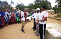 Four adult people stand infront of a crowd of Tanzanian school children holding malaria net materials.