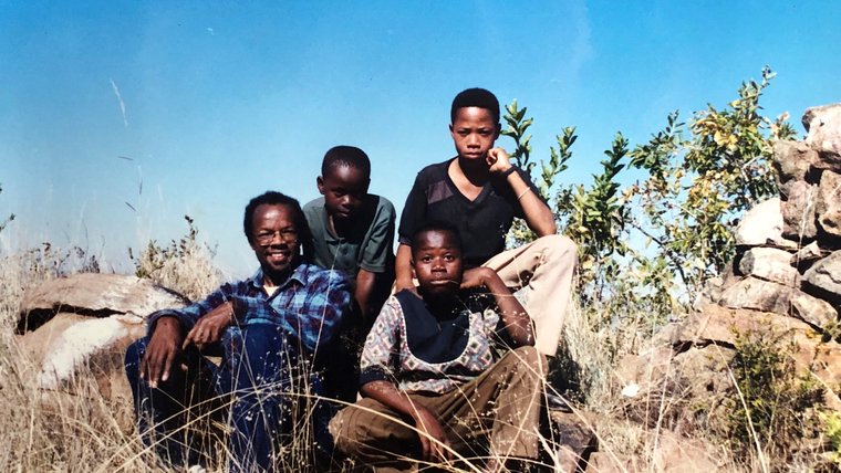 A middle aged black American man sits with two teenage South Africans on a hill outside.