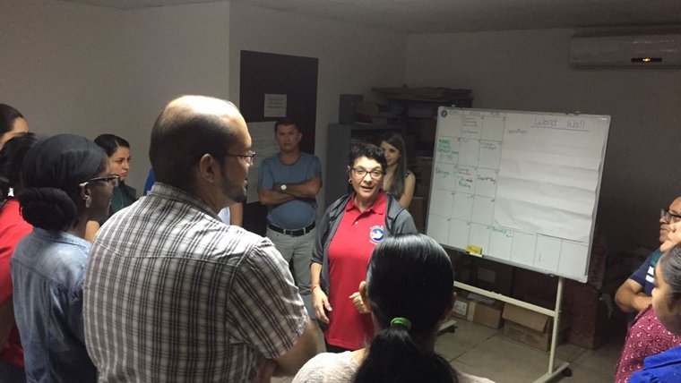 An American woman wearing a red Peace Corps polo gives a presentation in front of a group of her peers inside of an office.