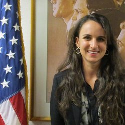 A young, white woman stands next to an American flag and smiles for a professional head shot..