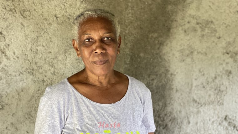 A older, black Jamaican woman stands in front of a grey wall and smiles.