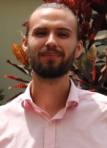 A bearded young male person with a pink shirt stands in front of a flower grove