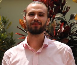 A bearded young male person with a pink shirt stands in front of a flower grove