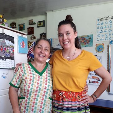 Two women stand in an office, their arms around each other's shoulders. They are smiling.