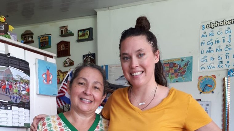 Two women stand in an office, their arms around each other's shoulders. They are smiling.