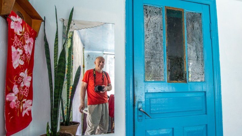 A man stands with his camera around his neck in front of a mirror. To the right is a bright blue door, to the left is a plant