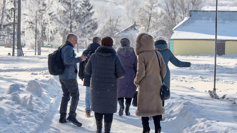 A group of people walk on a snowy road, their backs to the camera. There are trees and buildings in the background.