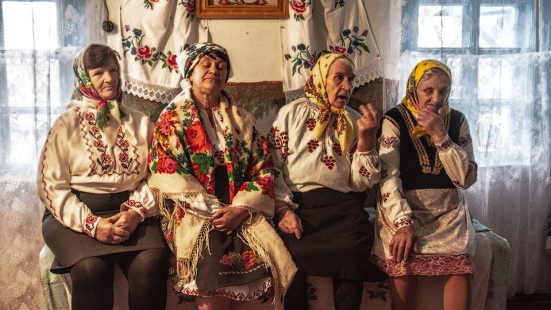 Four older Ukrainian women sit on a bench, wearing scarves on their heads and dressed in traditional clothing.