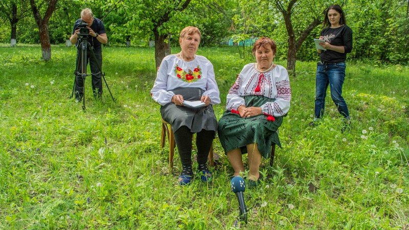 Two Ukrainian women dressed in traditional clothing sit on chairs in a green field. There is a camera crew behind them.