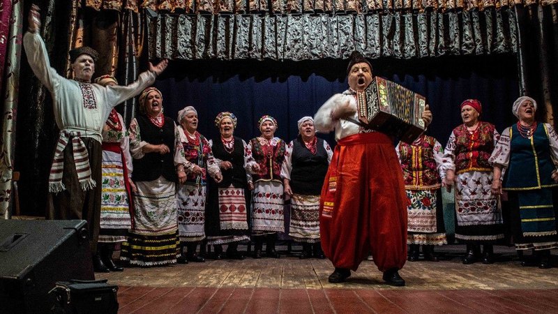 A man stands in the center of a stage playing an accordion and singing. Men and women in traditional clothing sing behind him