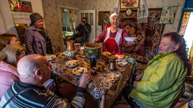 A group of Ukrainians sit around a table filled with food. They are in a colorful room.