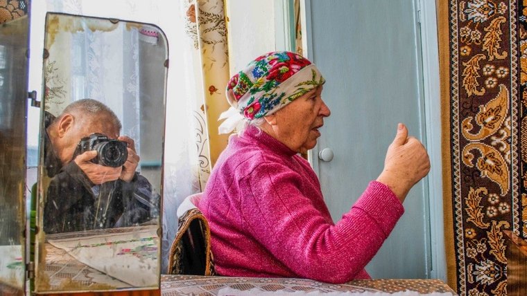 A woman sits amid an ornately decorated room, telling a story. Michael is reflected in a mirror taking her photo.