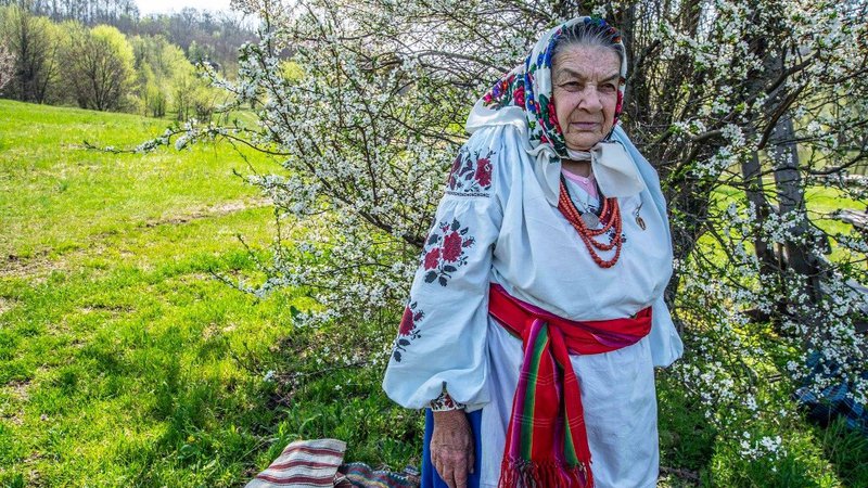 An older Ukrainian woman stands in traditional attire amid a green field and blooming tree.