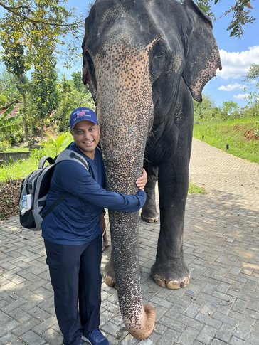 A man in an all blue outfit hugs an elephant's trunk.