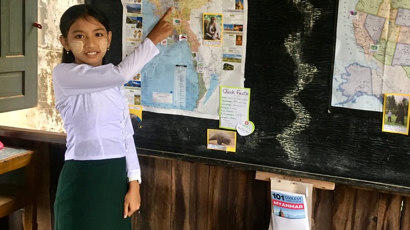 A girl stands and smiles while pointing to a map of Myanmar.