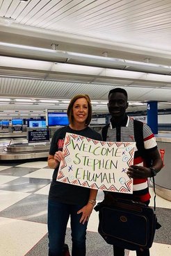 A female Returned Peace Corps Volunteer stands in an airport with her Ghanaian friend holding a "welcome Stephan" sign.
