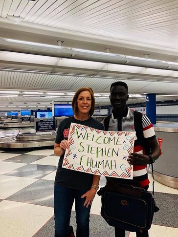 A female Returned Peace Corps Volunteer stands in an airport with her Ghanaian friend holding a "welcome Stephan" sign.