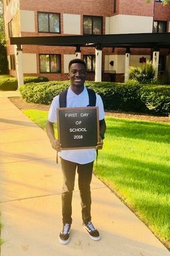 A Ghanaian young male stands outside smiling with a "first day of school" sign.