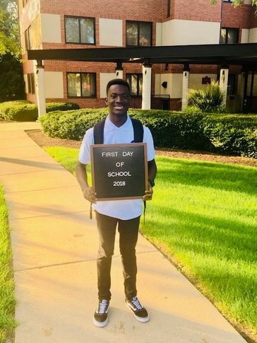 A Ghanaian young male stands outside smiling with a "first day of school" sign.