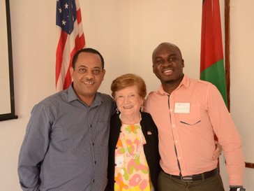 An older, white female Volunteer stands with a Malawian counterpart and another co-worker infront of an American flag and a M
