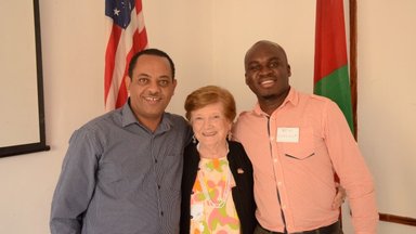 An older, white female Volunteer stands with a Malawian counterpart and another co-worker infront of an American flag and a M