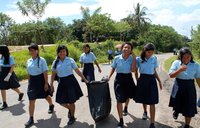 A group of girls in school uniforms walk down a street holding a trash bag, smiling while they participate in a community cle