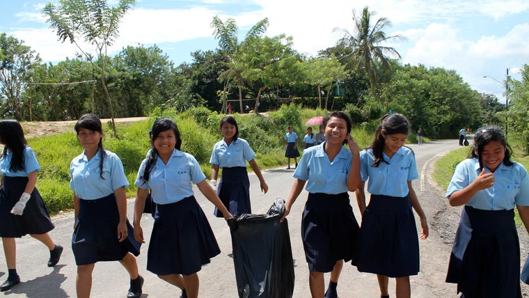 A group of girls in school uniforms walk down a street holding a trash bag, smiling while they participate in a community cle