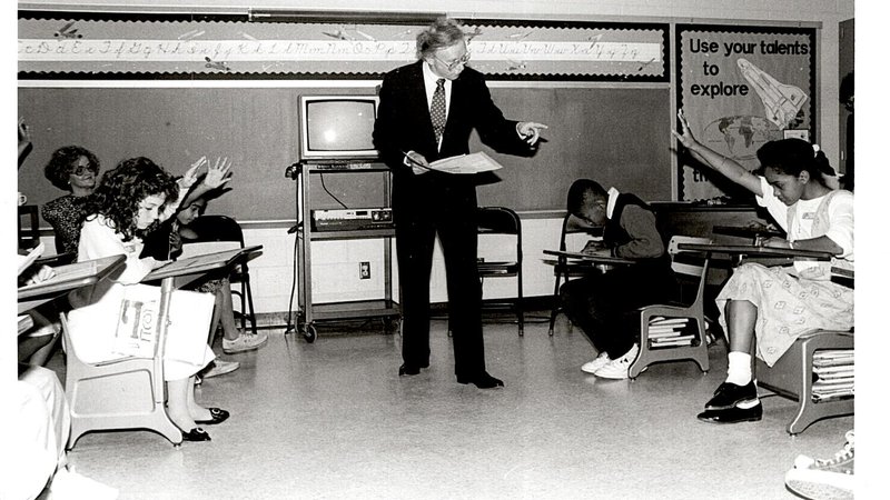 In a black and white photo, former Peace Corps Director Paul D. Coverdell stands in a classroom with young children, pointing