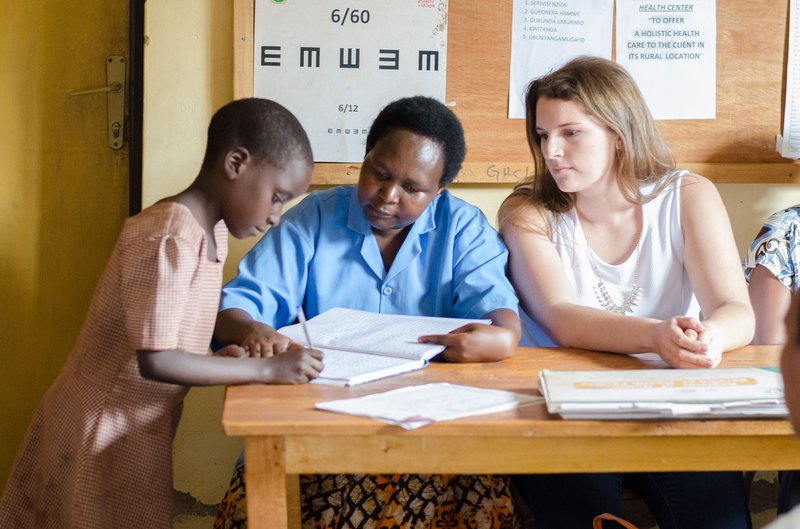 PCV Caroline and her counterpart sit at a desk as a child writes the family's name on the log in sheet.
