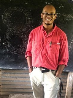 A Black American man in dress clothes stands in front of a chalkboard