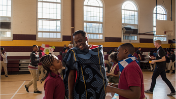 Young Black students look at and listen to a Black male dressed in traditional clothing.