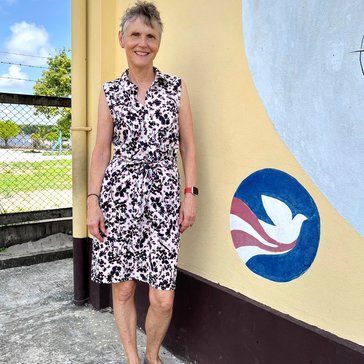 Female stands near painted wall (Peace Corps logo) in cotton sleeveless, knee length dress and flats