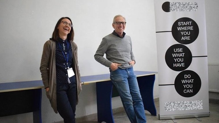 A man and woman stand next to each other, both leaning back on a table behind them. They are smiling and laughing, and beside