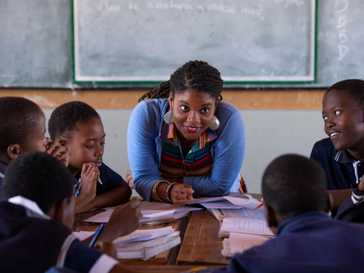 A Peace Corps Volunteer in South Africa leans over the front of a table, smiling as she talks to her young students.