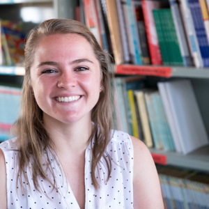 A Caucasian female Peace Corps Volunteer smiles in front of a bookshelf.