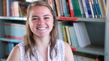 A Caucasian female Peace Corps Volunteer smiles in front of a bookshelf.