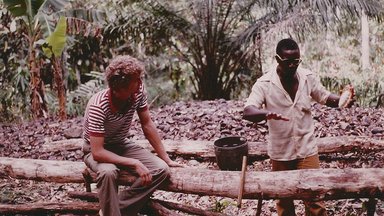 a white man sits on a fallen tree trunk, listening to a black man who holds a cocoa pod