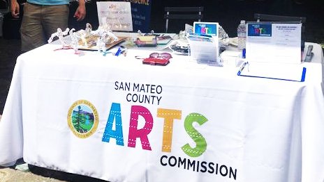 A booth at an art fair with brightly colored letters on a white tablecloth that reads San Mateo Arts and a white man in back