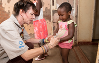 An older Peace Corps Volunteer shows a young girl how to use hand sanitizer. They're facing each other and their profiles are