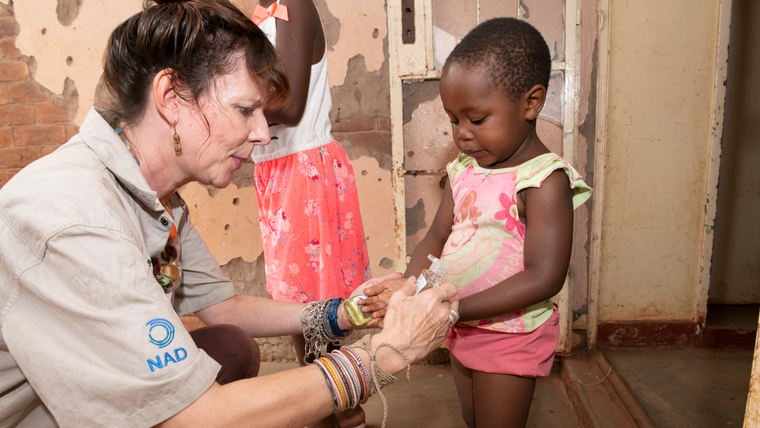 An older Peace Corps Volunteer shows a young girl how to use hand sanitizer. They're facing each other and their profiles are