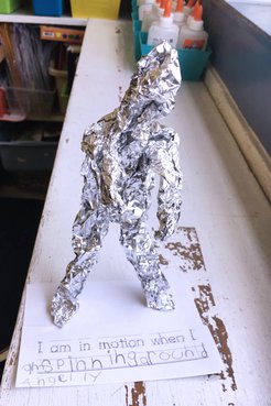 A close up of a foil sculpture that looks like a lumpy man made by a Jamaican primary school student on top of a table
