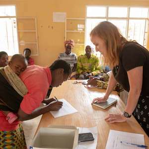 A red-haired Peace Corps Volunteer stands across the table from a mother in Rwanda. The mom has a baby tied to her back.