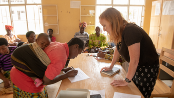 A red-haired Peace Corps Volunteer stands across the table from a mother in Rwanda. The mom has a baby tied to her back.
