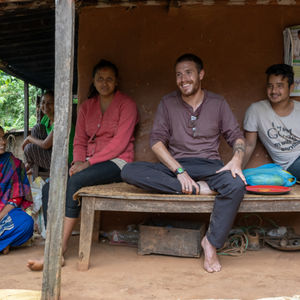 A male Peace Corps Volunteer sits on a bench outside between two Nepali people. They are all barefoot.