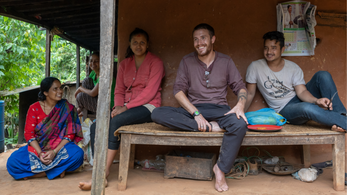 A male Peace Corps Volunteer sits on a bench outside between two Nepali people. They are all barefoot.