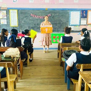 Female at the front of class (facing camera) holding cardboard poster with information about reproductive health. Female students are sitting in benches backing camera.