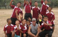 A male Volunteer and a group of boys dressed in baseball uniforms, smile and pose for a photo on a field.