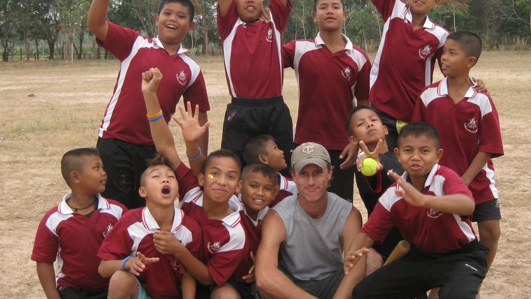 A male Volunteer and a group of boys dressed in baseball uniforms, smile and pose for a photo on a field.