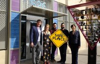 Five coworkers pose for a picture outside of their office building. They are holding a yellow sign that says "Safe Place"