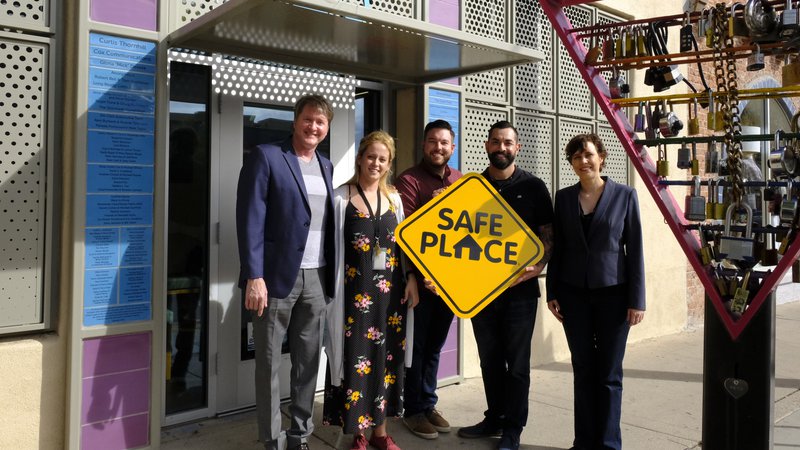 Five coworkers pose for a picture outside of their office building. They are holding a yellow sign that says "Safe Place"